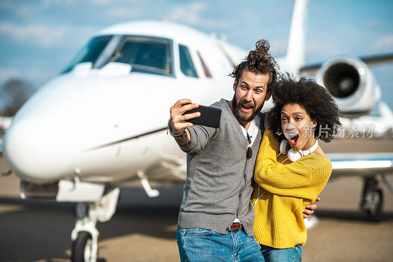 一对富有的年轻夫妇正在机场停机坪上的一架私人飞机前用手机自拍