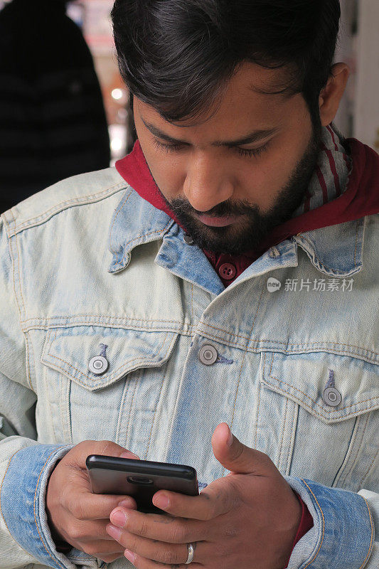 在印度德里，英俊的印度男子用手机看屏幕发短信，查看社交媒体和互联网，年轻的印度男子用手机交流，城市背景，连帽衫青年牛仔夹克
