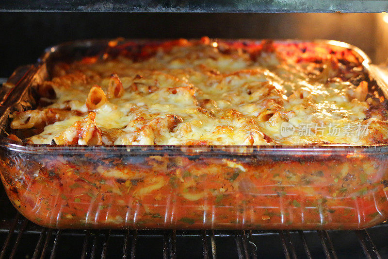 这是一个自制的意大利面食烘焙食谱，放在方形玻璃盘子里，用意大利通心粉管夹着浓郁的肉酱番茄酱、洋葱、香草、新鲜番茄和切碎的罗勒叶，在热的厨房烤箱里烤着，帕尔马干酪和切达干酪融化在上面