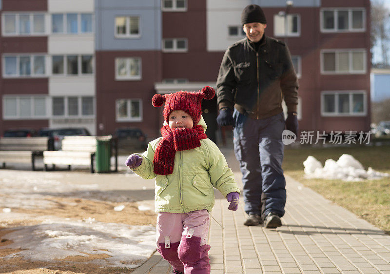 2岁的小女孩在室外人行道上跑步，而他70岁的爷爷则走在后面。