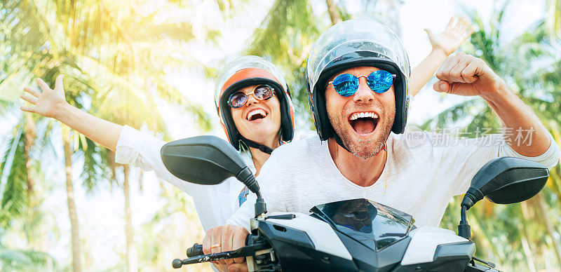 在泰国苏梅岛棕榈树下的热带度假中，一对快乐微笑的旅行者戴着安全帽骑着摩托车
