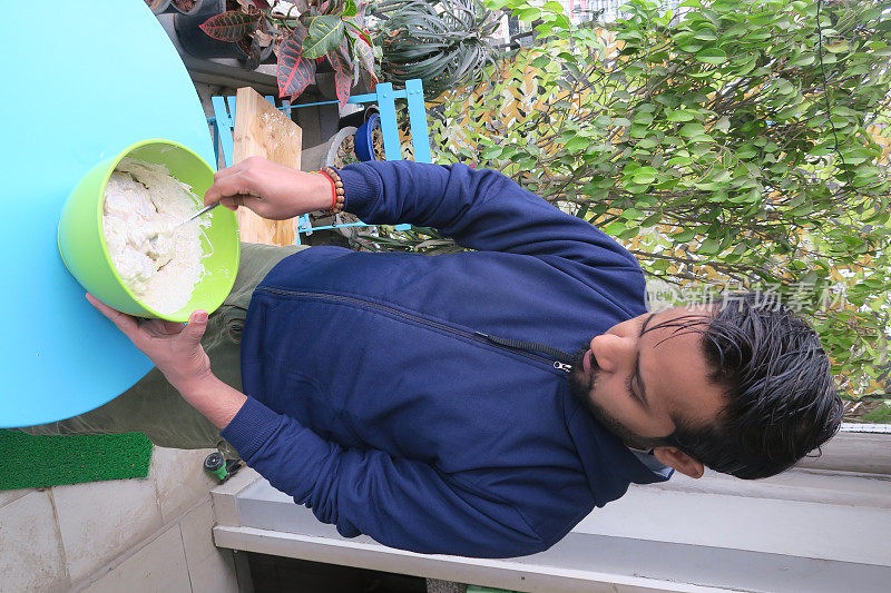 英俊的印度男子在一碗奶酪和酸奶腌制的生鸡肉，鸡胸肉准备在煎锅和烧烤串，英俊的男子穿着蓝色连帽衫站在阳台上与绿树的背景