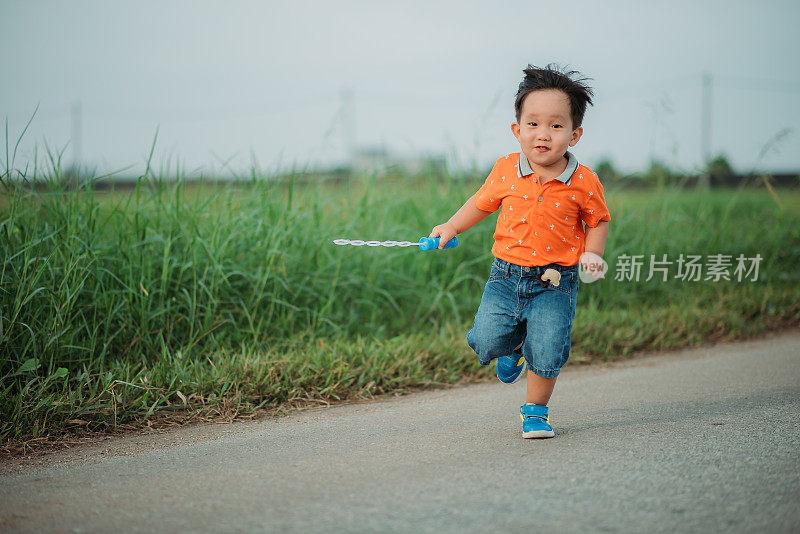 亚洲华人微笑男孩花闲暇时间，手拿泡泡棒，在村里的街道上跑步