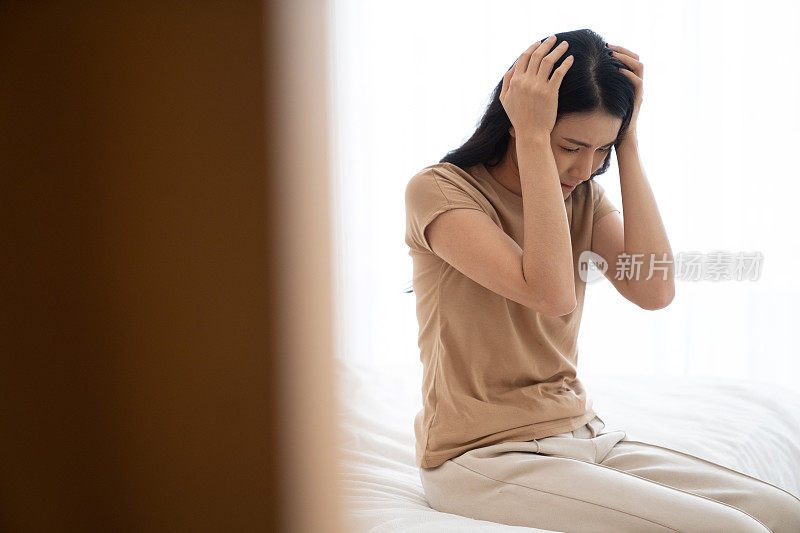 年轻的亚洲女人坐在她房间的床上，感到压力和沮丧。