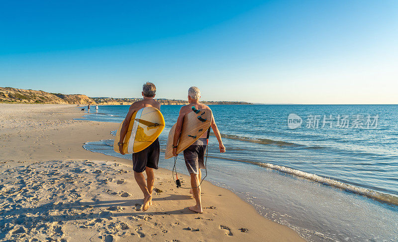 两个成熟的男人带着冲浪板在美丽的海滩上散步，享受着天堂和退休生活。有吸引力的老年人朋友有乐趣的冲浪。在真正积极健康的人身上。