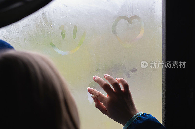少女或年轻女子用手在雾气蒙蒙的窗户上作画。手指在湿玻璃上画出了心、窗和雨