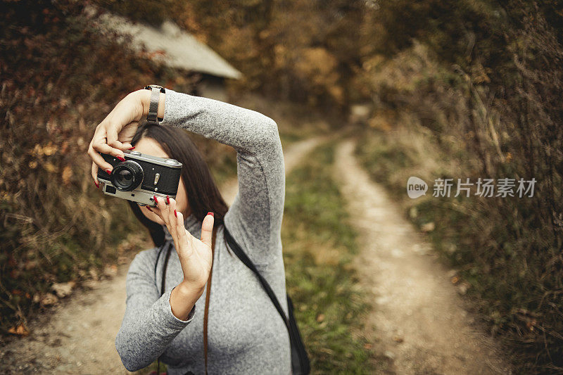 在大自然中摄影——快乐的年轻女子微笑着享受大自然，走过画着秋天颜色的森林，戴着复古的相机拍摄树叶