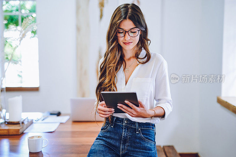 照片中，一位迷人的年轻女商人独自坐在她的家庭办公室的办公桌上，使用着数码平板电脑