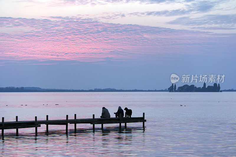 日落时分，夫妇和狗坐在湖边的码头上(XL)