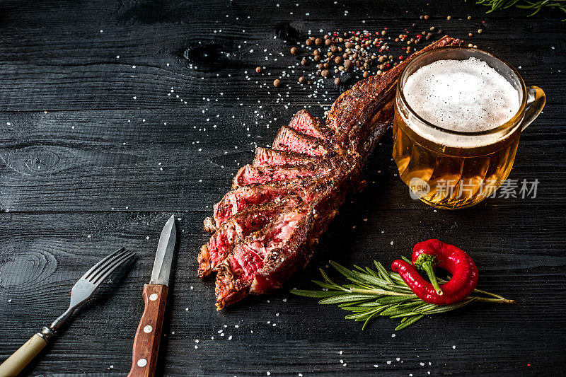 烧烤干燥的陈年牛肉肋与香料，蔬菜和一杯淡啤酒近距离上的黑色木制背景