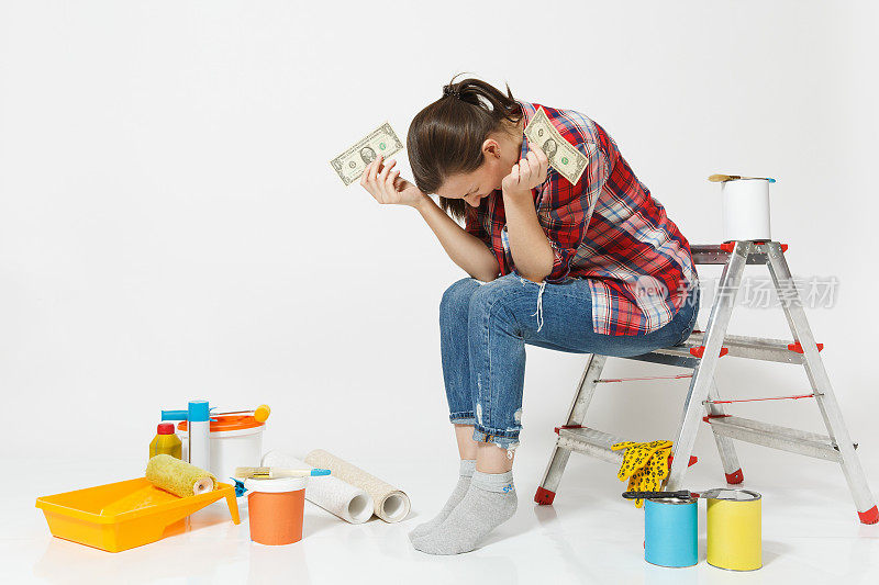 心烦意乱的女性紧握着美元，现金很少的钱，坐在梯子上用仪器装修公寓孤立在白色的背景。墙纸、粘漆配件工具。修复的概念