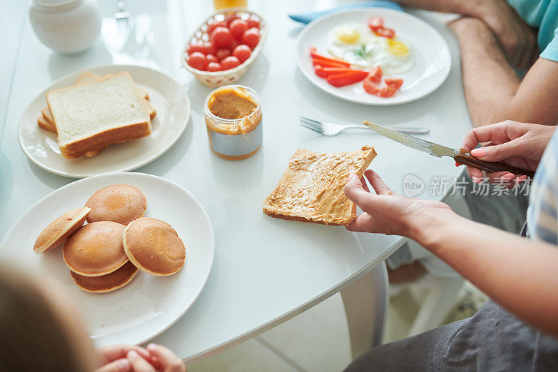 特写镜头:一个面目全非的女人正在为家庭早餐准备花生酱吐司，她站在一张有煎饼和煎蛋的桌子前