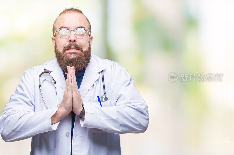 年轻的白种人医生男子身穿医疗白大褂在孤立的背景乞求和祈祷与希望的表情在脸上非常情绪化和担心。请求宽恕。宗教的概念。