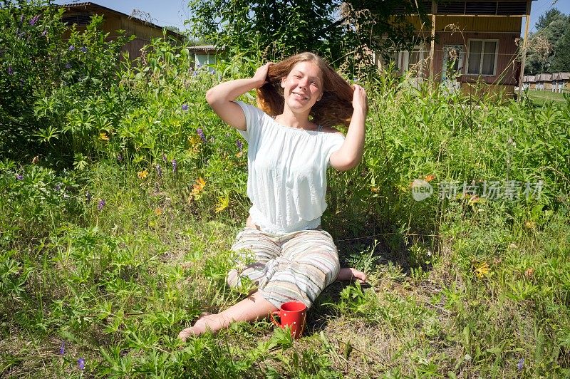 快乐的女孩坐在草地上，用一杯红色的咖啡在露营的背景，夏天的早晨。