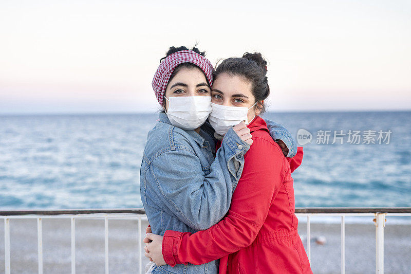 两个戴着防护口罩的女朋友在海滩上互相拥抱，这是一种新的常态