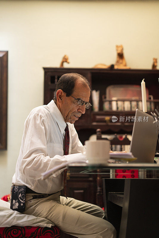 一个成年男子坐在家里的餐厅里，一边用笔记本电脑一边上虚拟课程