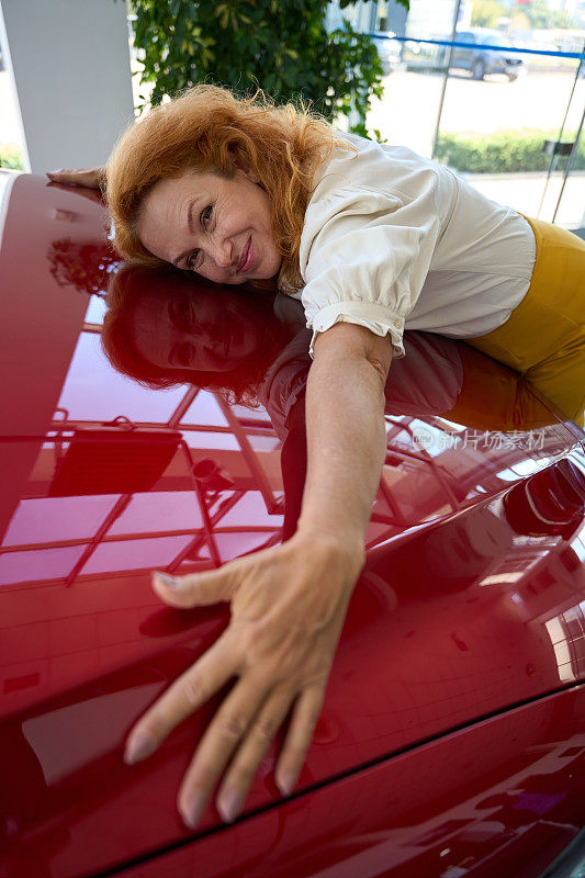 快乐的女人在汽车陈列室拥抱汽车引擎盖