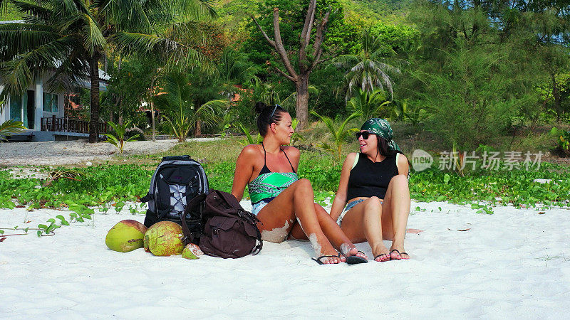 在泰国苏梅岛，两名白人美女穿着比基尼，背着包坐在沙滩上