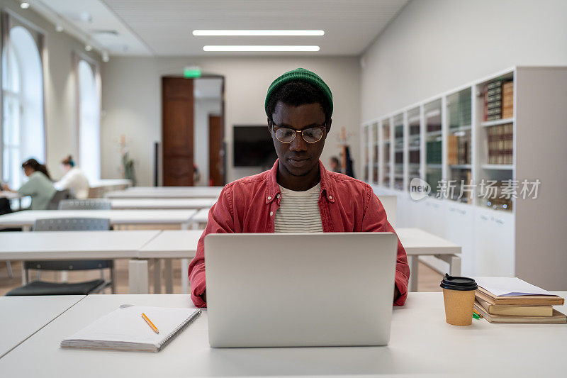 严肃沉思的黑人学生男子戴眼镜坐在桌子上使用笔记本电脑在大学讲座教室