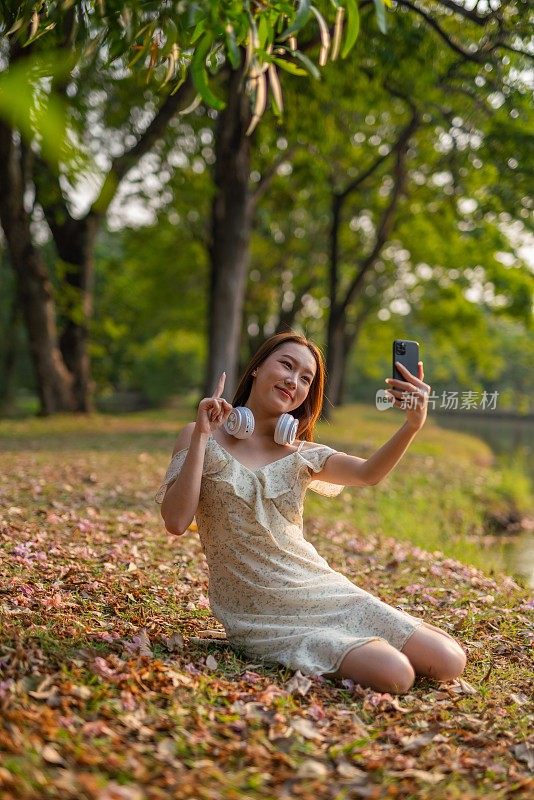 年轻的亚洲女性在公园户外自拍。带着耳机的美女在花园里享受大自然