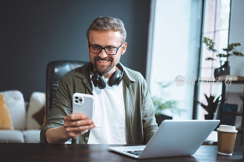 一个快乐微笑的男人坐在阁楼办公室里，穿着休闲装，一边用笔记本电脑工作，一边用手机发短信。