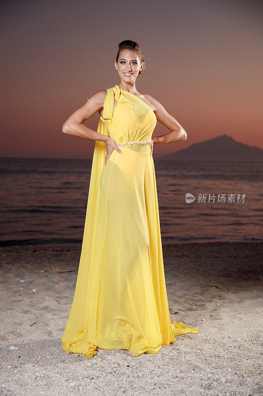 美丽的年轻女子穿着黄色无袖长裙，双手叉腰站在沙滩上