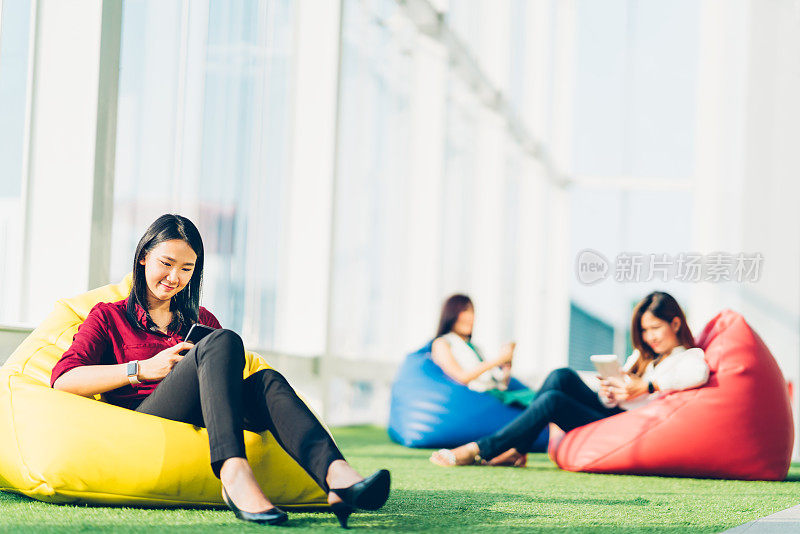 一群使用智能手机的亚洲大学生或商业同事，一起坐在现代办公室或大学校园里。社交媒体网络，手机生活方式，或通讯技术概念