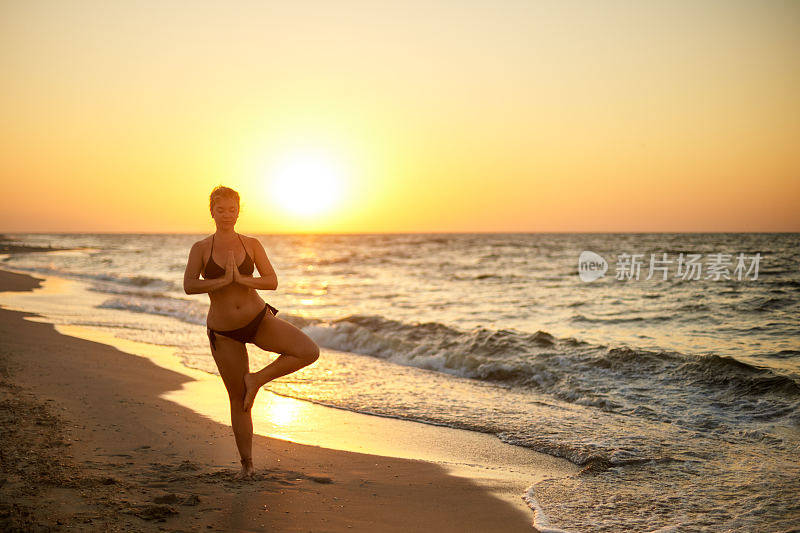 真实的女人在做瑜伽的泳衣在早上的海滩上。真实的未经修饰的女孩剪影在树的姿势在日出海边体式。身体积极的女性在日落训练
