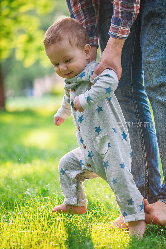 在夏天的草地上，小男孩很高兴他的父亲扶着他迈出了第一步
