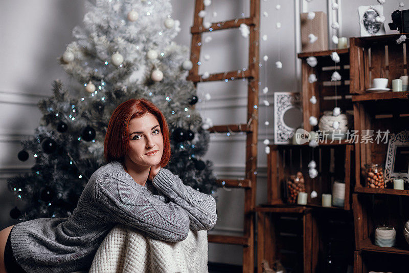 时尚室内照片美丽的年轻女子红头发和迷人的微笑，穿着舒适的针织羊毛衫，摆姿势与圣诞树和巧克力蛋糕与星号的礼物