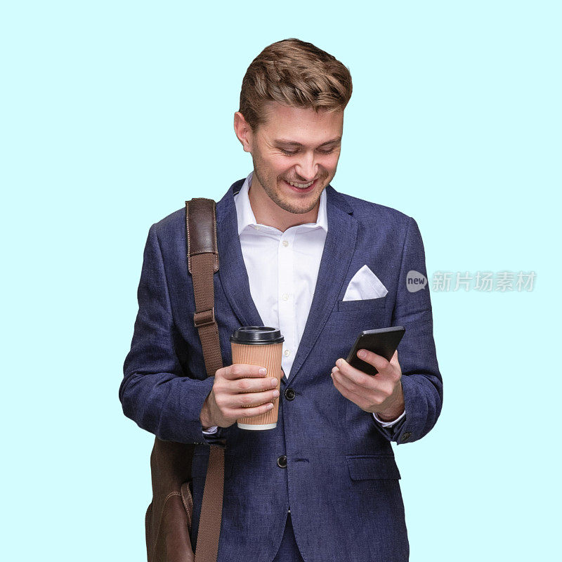 站在蓝色背景的白人年轻男性商人面前，背着小提包，拿着咖啡杯，使用智能手机