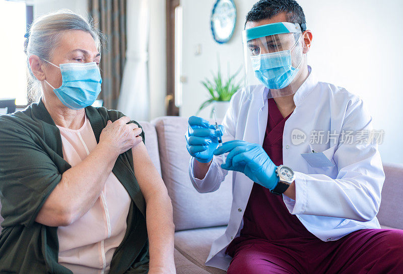 在冠状病毒爆发期间，佩戴防护口罩坐在家中并给一名老年妇女注射的男护士。老年患者在家接种疫苗。冠状病毒疫苗接种