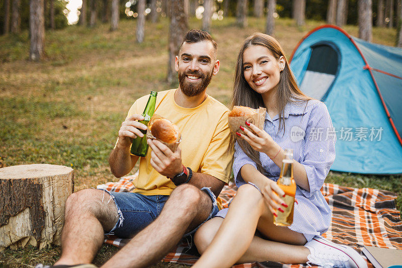 快乐的年轻夫妇有露营晚餐，喝啤酒和吃汉堡在森林