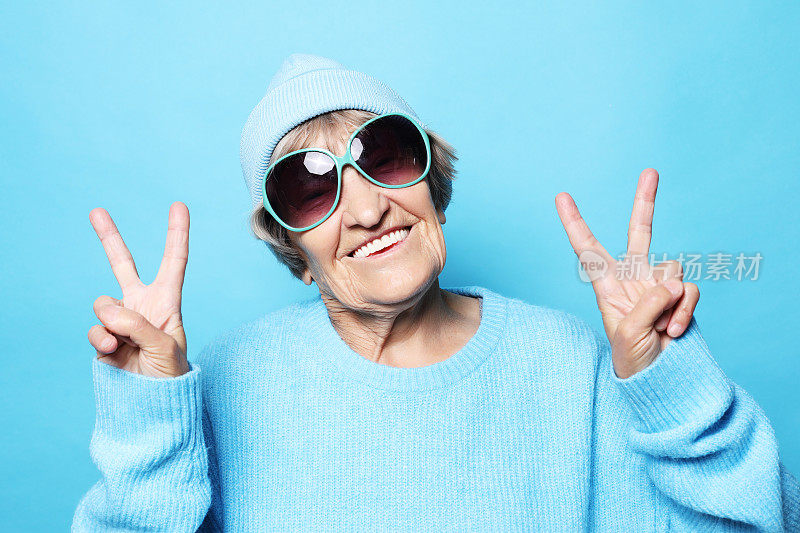 生活方式，情感和人的概念:有趣的老妇人穿着蓝色毛衣，帽子和太阳镜显示胜利的标志
