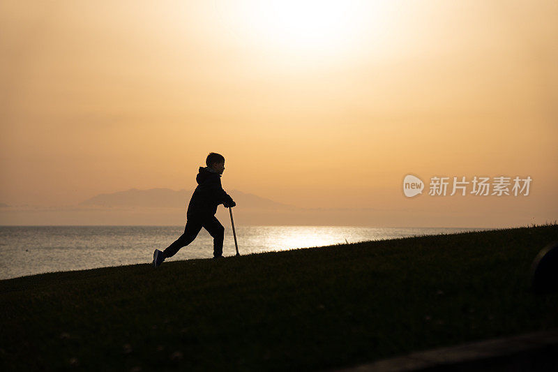 日落时分，小男孩推着他的踏板车走在海边的小山上