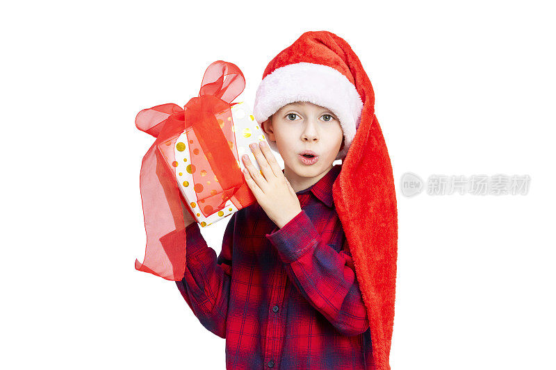 惊讶的男孩睁大了眼睛和嘴巴，拿着和摇晃着一个有红色蝴蝶结的礼品盒。孩子戴着圣诞帽，穿着睡衣，带着礼物，看着白色背景的相机。