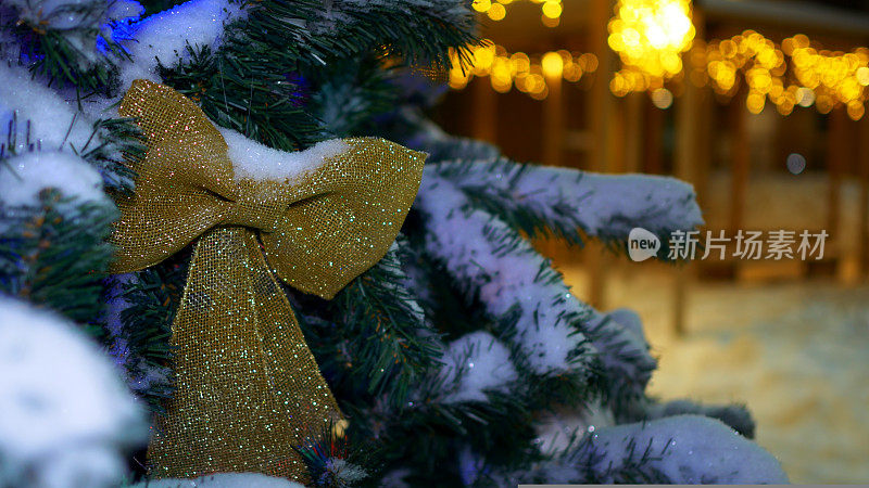 圣诞节的气氛在晚上的街上，黄色的花环装饰着房子，一棵装饰好的圣诞树上装饰着金色的蝴蝶结，上面覆盖着新雪。圣诞夜。