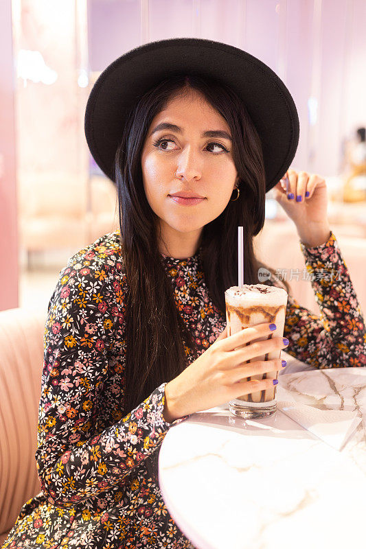 一位戴着帽子的年轻女子坐在餐厅里，手里拿着一杯冰沙