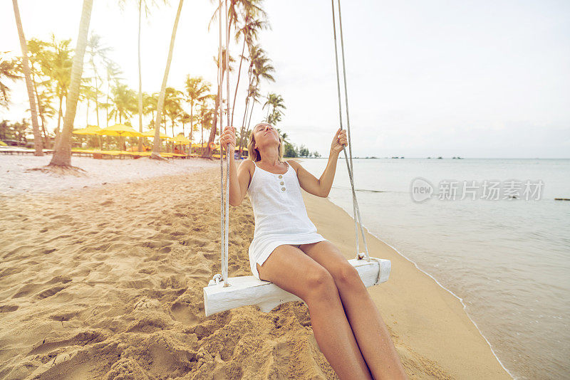 俏皮的年轻女子在热带海滩使用秋千在日出