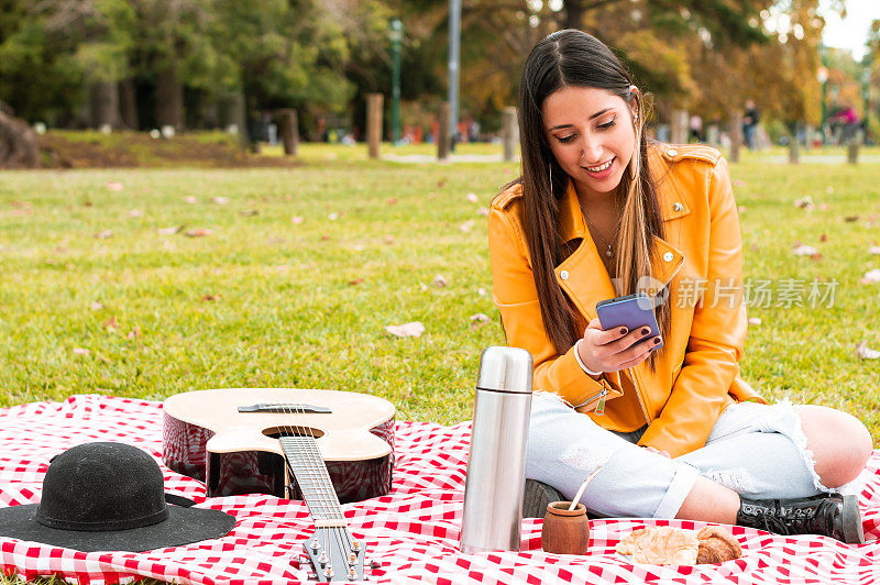 放松的年轻拉丁女子在公园野餐，坐在娱乐查看她的手机
