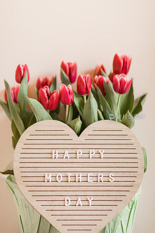 一个心形的字母板，上面写着“母亲节快乐”，明亮的粉红色郁金香盛开，郁郁葱葱的绿色茎和叶子在它后面，在明亮的自然日光下拍摄，中性的米色背景与复制空间