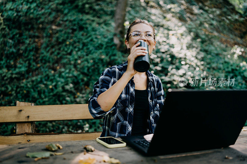 一位年轻女子将手提电脑工作与在自然环境中喝水相结合