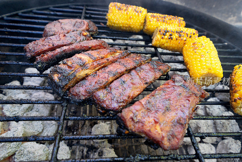 烧烤婴儿背部猪肉排骨在一个火热的烧烤与烧焦的玉米棒子在背景