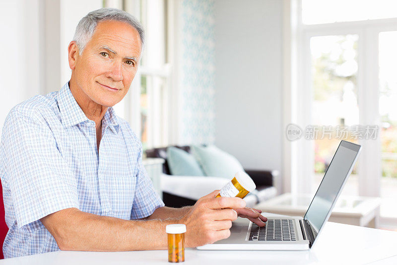 拿着药瓶在餐桌上使用笔记本电脑的老人。
