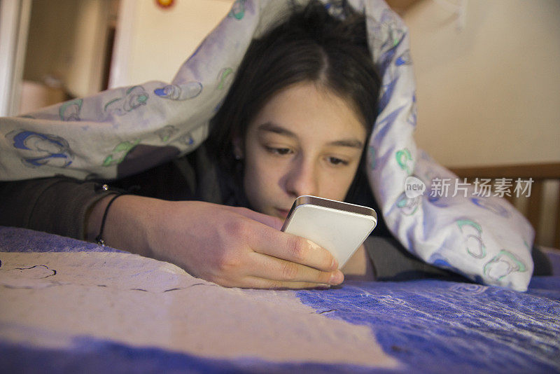 在床上躺在毯子下使用手机的青少年