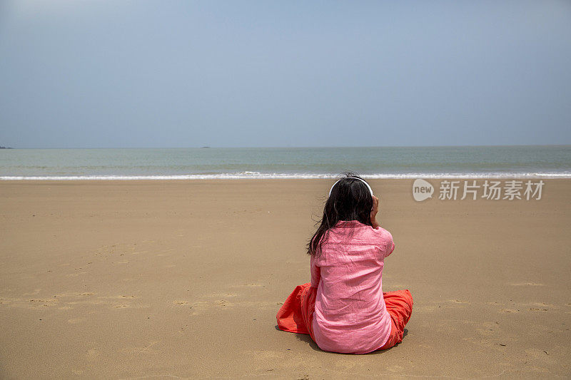 女孩坐在海滩上，听着音乐，面朝大海