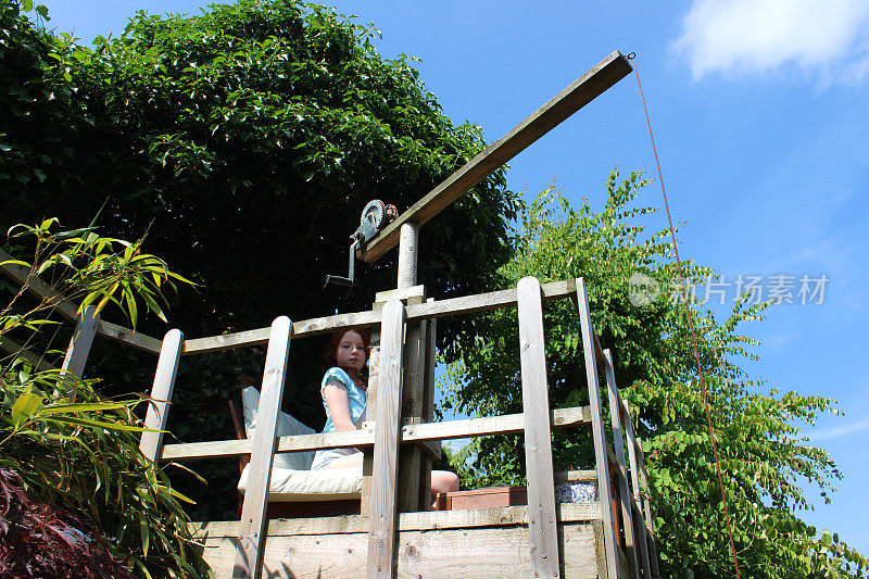 女孩坐在树屋的平台上，木制起重机玩具，尖桩栅栏
