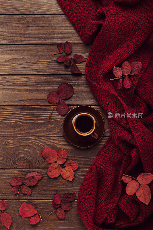 编织的紫红色围巾与秋天的叶子和一杯咖啡在一个深色的木制背景。