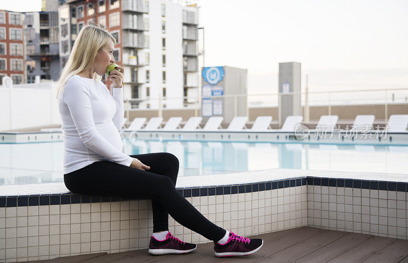 这名年轻的孕妇是25岁的白人，在健身完后，她在屋顶游泳池附近休息时正在吃苹果。