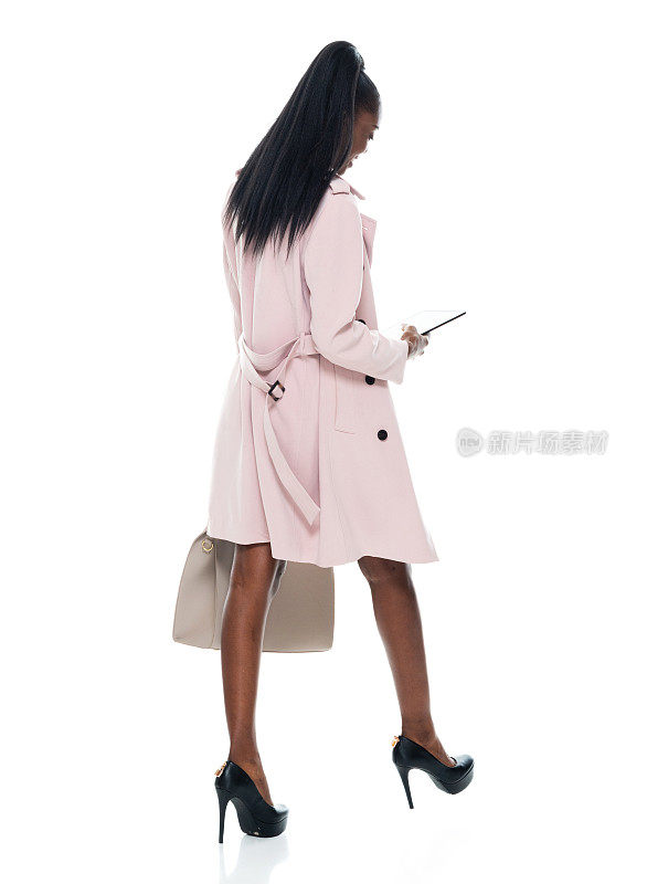 非洲裔女性走在白色背景前，穿着风衣，拿着钱包，使用数码平板电脑